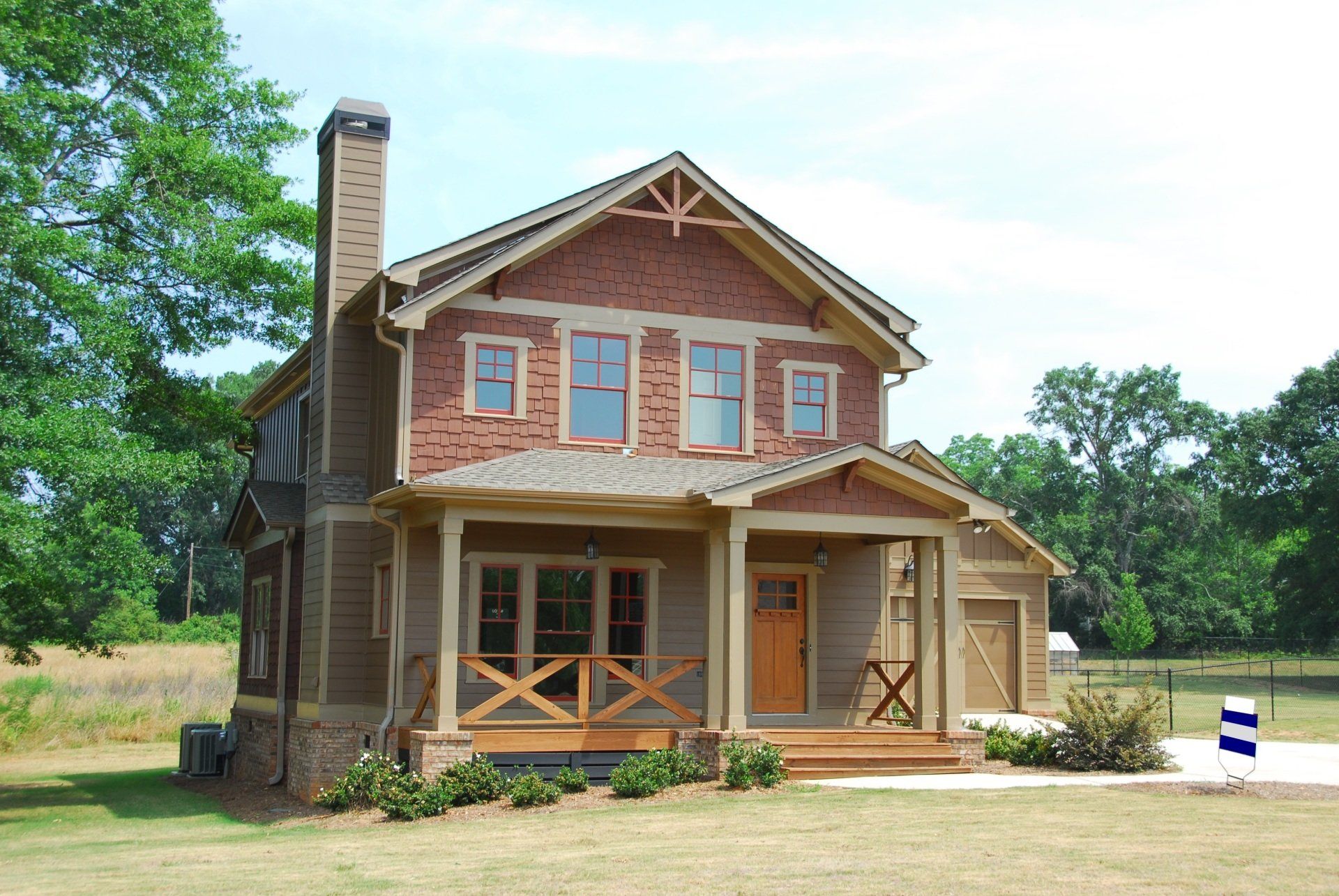 Two-story brick house with a wraparound porch and chimney in a grassy, wooded setting