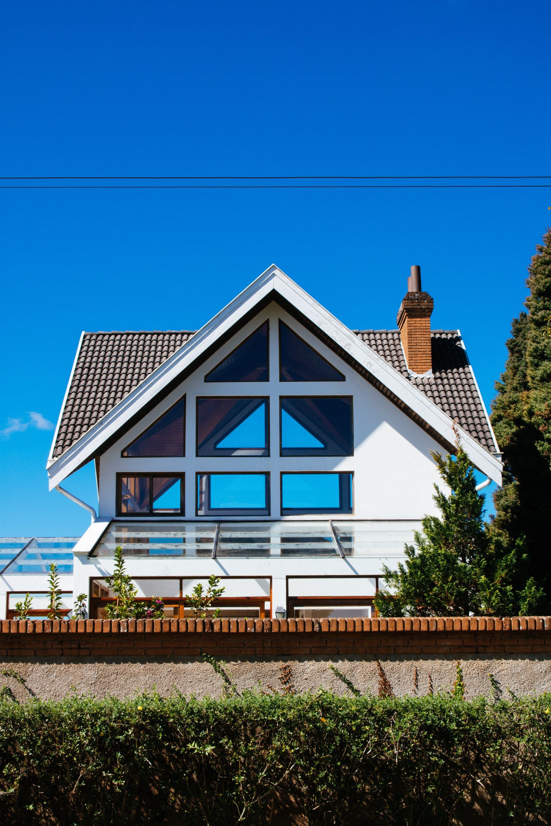 White house with blue windows and a tiled roof under a clear blue sky, behind a hedge and brick wall